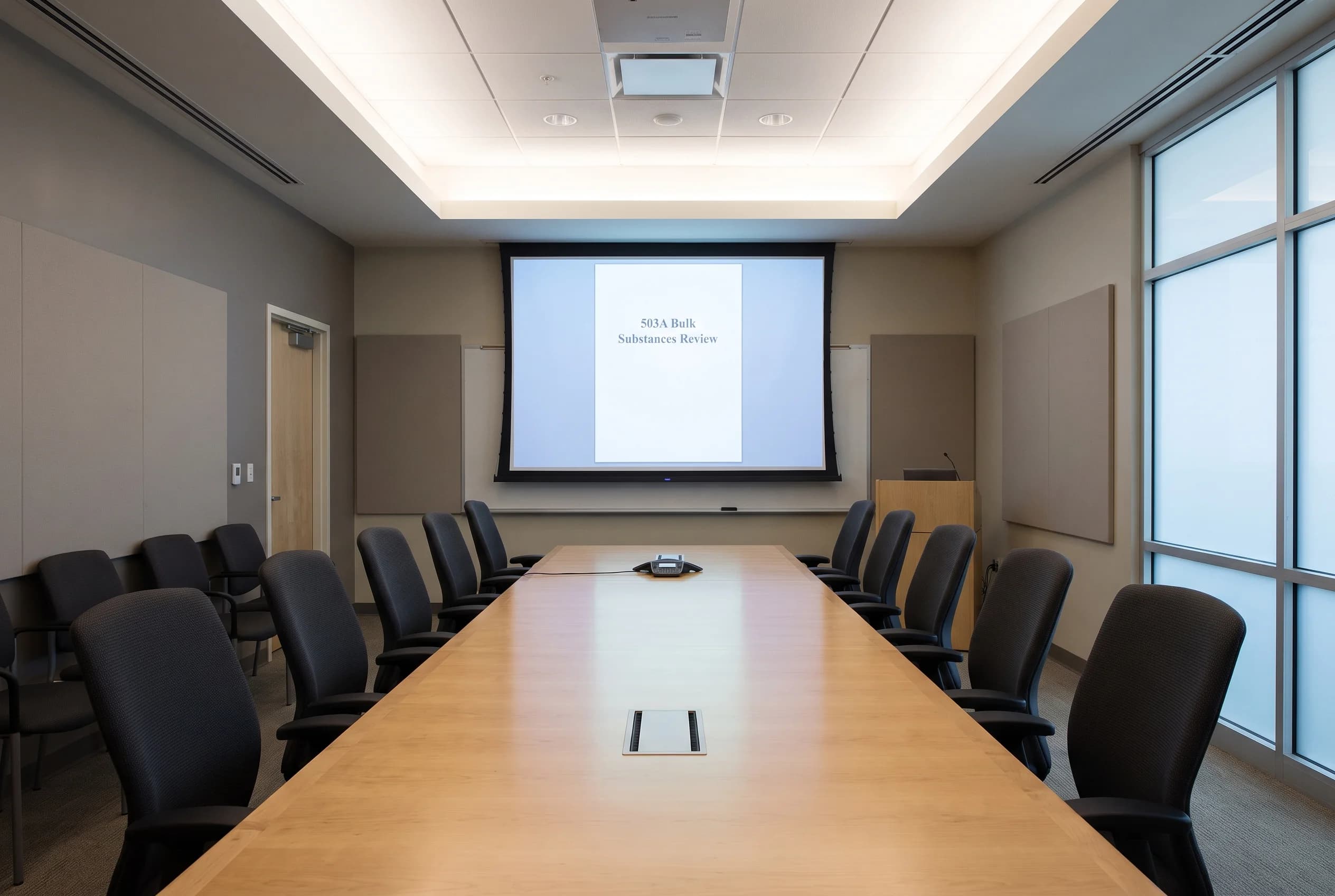 An empty government advisory meeting room with a long rectangular table, empty chairs, and a projection screen displaying a generic agenda document