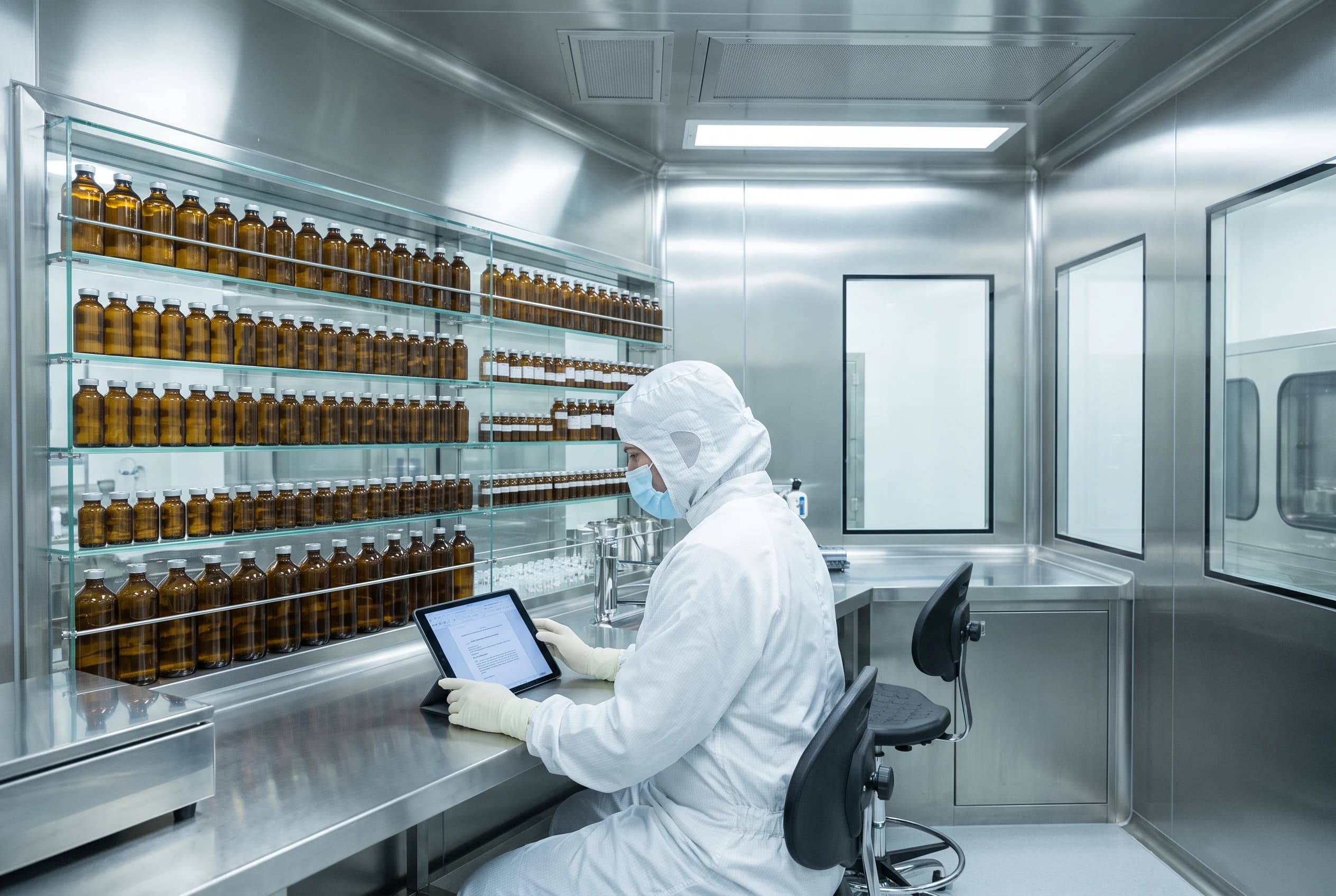 Clean-room-gowned operator reviewing a document on a tablet beside shelves of amber pharmaceutical bottles in a sterile compounding room