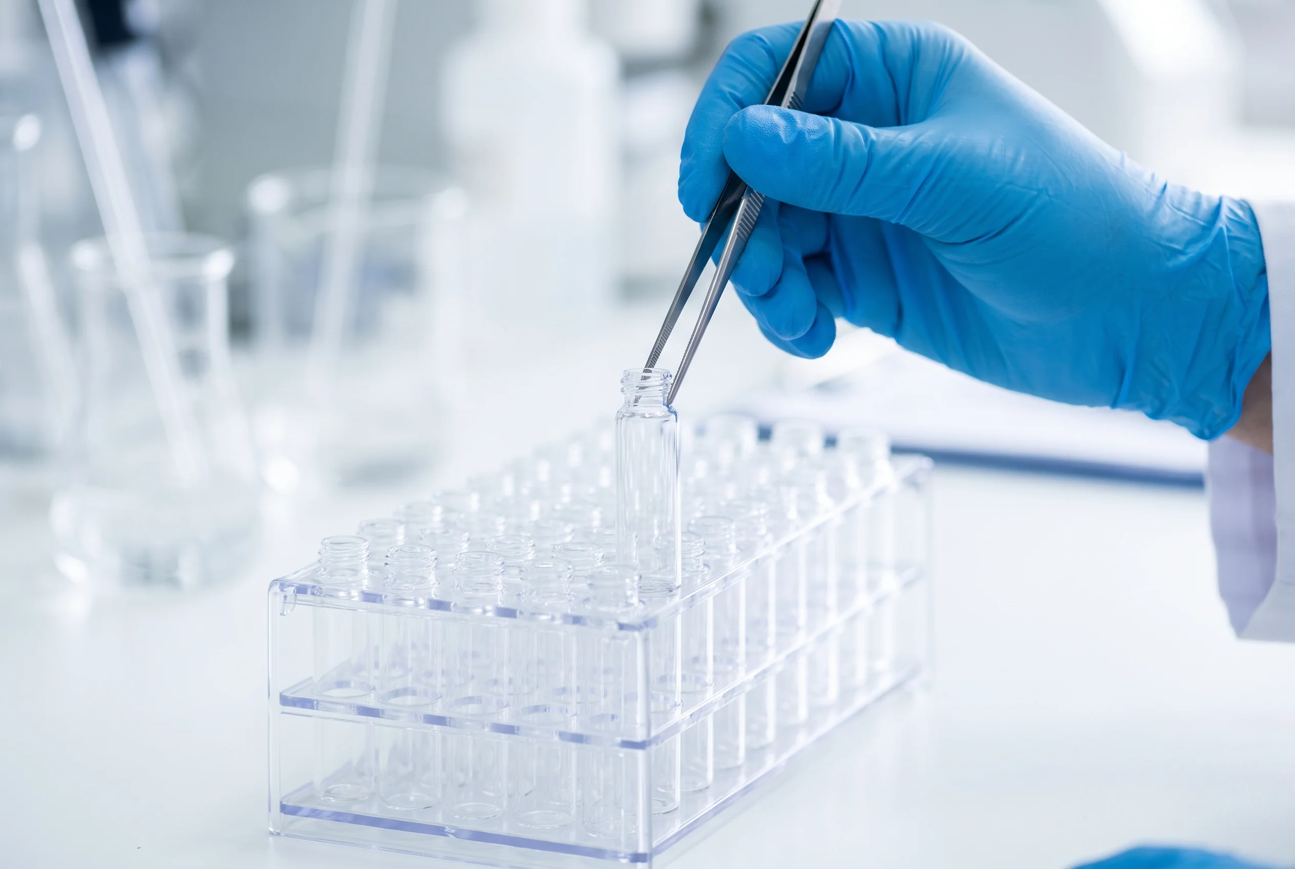 Blue-gloved researcher using tweezers to place a small clear unlabeled glass vial into a laboratory sample rack